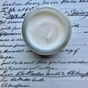 May include: A small glass jar filled with a white, creamy substance. The jar is sitting on a piece of paper with handwritten text that includes the words "Acid Salicylic", "Syr", "Belladonna", and "Chloroform".