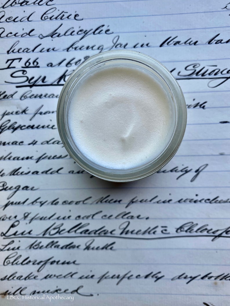 May include: A small glass jar filled with a white, creamy substance. The jar is sitting on a piece of paper with handwritten text that includes the words "Acid Salicylic", "Syr", "Belladonna", and "Chloroform".
