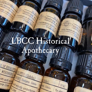 May include: A collection of small amber glass bottles with black caps, labeled with the text "LBCC Historical Apothecary". The bottles contain essential oils, including "Oil of Eucalyptus (Globulus)" and "Beaver Dam Wisconsin".