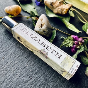 May include: A glass bottle with a silver cap and a label that reads "Elizabeth" with a small illustration of a bird and a tree. The bottle is sitting on a dark surface with small stones and green leaves.