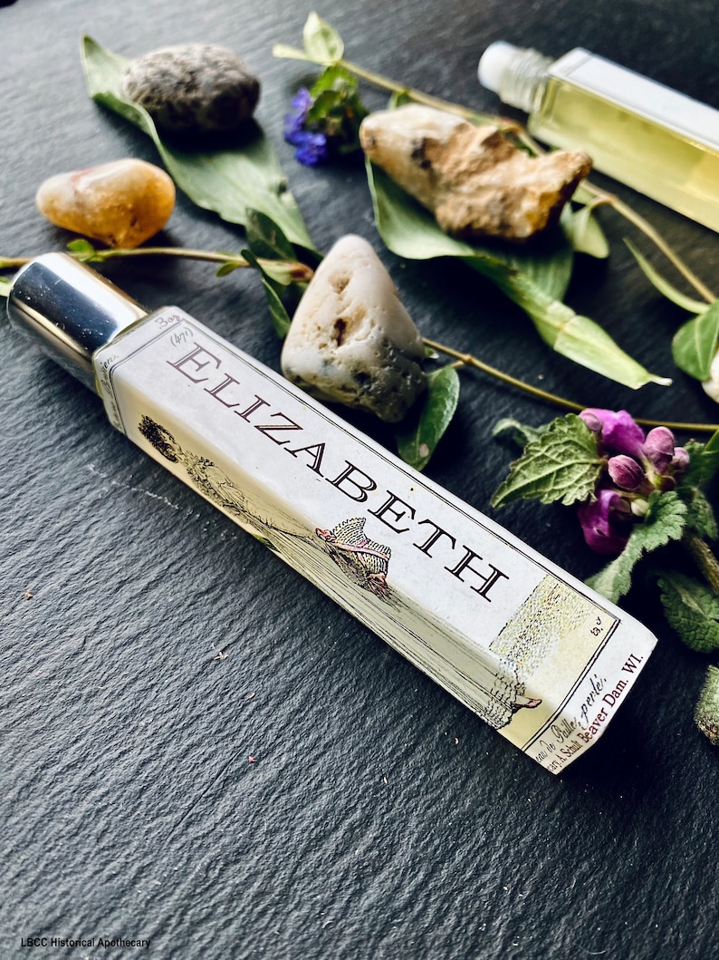 May include: A glass bottle with a silver cap and a label that reads "Elizabeth" with a small illustration of a bird and a tree. The bottle is sitting on a dark surface with small stones and green leaves.