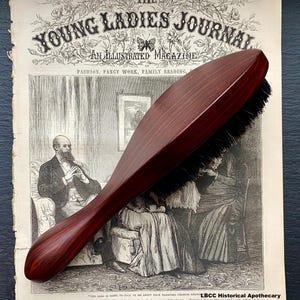 May include: A dark red wooden hairbrush with natural boar bristles. The brush is oval shaped and has a slightly curved handle. The brush is lying on top of an old magazine.