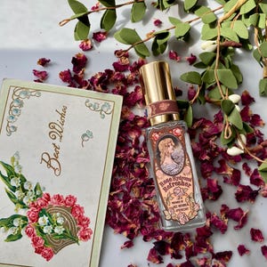 K&ouml;nnte beinhalten: Eine Glasflasche mit Rosen-Hydrolat-Erfrischer, hergestellt von LBCC Historical Apothecary. Die Flasche hat einen goldenen Verschluss und ein Etikett im Vintage-Stil mit einer Frau in einem Kleid und einem floralen Design. Die Flasche ist von getrockneten Rosenbl&uuml;ten und gr&uuml;nen Bl&auml;ttern umgeben.