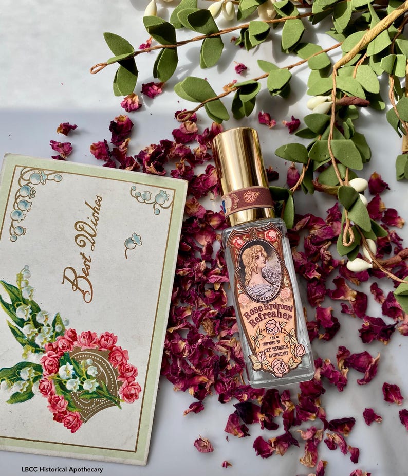 K&ouml;nnte beinhalten: Eine Glasflasche mit Rosen-Hydrolat-Erfrischer, hergestellt von LBCC Historical Apothecary. Die Flasche hat einen goldenen Verschluss und ein Etikett im Vintage-Stil mit einer Frau in einem Kleid und einem floralen Design. Die Flasche ist von getrockneten Rosenbl&uuml;ten und gr&uuml;nen Bl&auml;ttern umgeben.