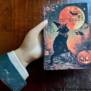 May include: A black cat wearing a witch's hat sits on a tree stump in front of a large orange moon. The moon has the words "My Notebook" written on it. The background is a dark forest with fall leaves and a jack-o-lantern.