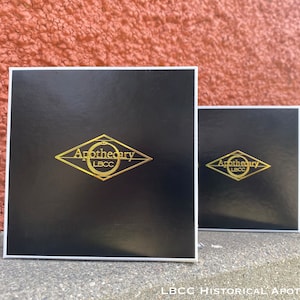 May include: Three black boxes with white borders and gold lettering. The boxes have the text "Apothecary LBCC" on them. The boxes are arranged in a row, with the largest box in the front and the smallest box in the back.
