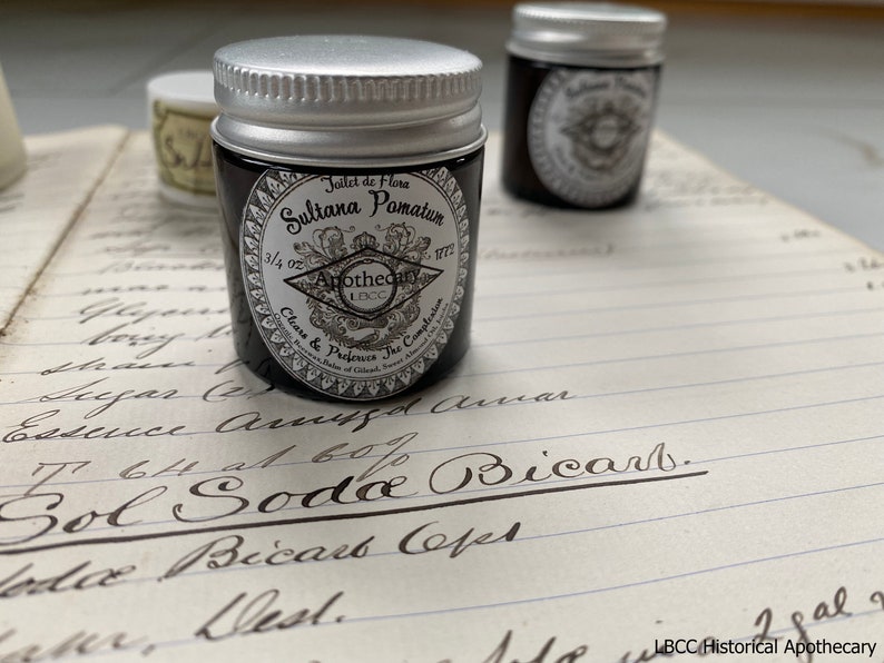 May include: A small, black jar with a silver lid. The jar is labeled "Joilet de Flora Sultana Pomatum 3/4 oz Apothecary 18CC Clears & Perfects The Complexion"