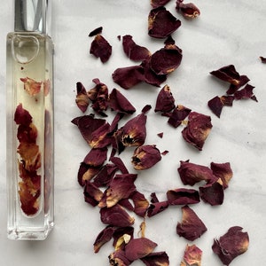 K&ouml;nnte beinhalten: Eine durchsichtige Glasflasche gef&uuml;llt mit getrockneten Rosenbl&uuml;ten und einer klaren Fl&uuml;ssigkeit. Die Flasche hat einen silbernen Verschluss. Die Flasche ist von getrockneten Rosenbl&uuml;ten auf wei&szlig;em Hintergrund umgeben.