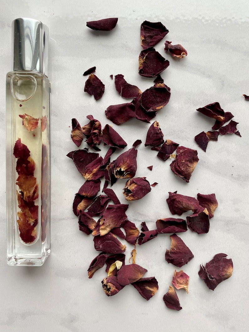 K&ouml;nnte beinhalten: Eine durchsichtige Glasflasche gef&uuml;llt mit getrockneten Rosenbl&uuml;ten und einer klaren Fl&uuml;ssigkeit. Die Flasche hat einen silbernen Verschluss. Die Flasche ist von getrockneten Rosenbl&uuml;ten auf wei&szlig;em Hintergrund umgeben.