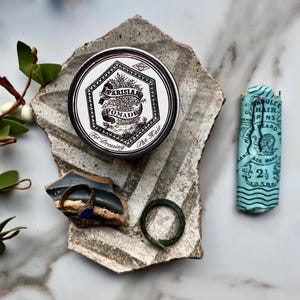 May include: A vintage tin of Parisian Pomade for dressing the hair. The tin is silver with a white label and black lettering. The label features a stylized image of a plant and the text "Parisian Pomade" and "For Dressing The Hair".