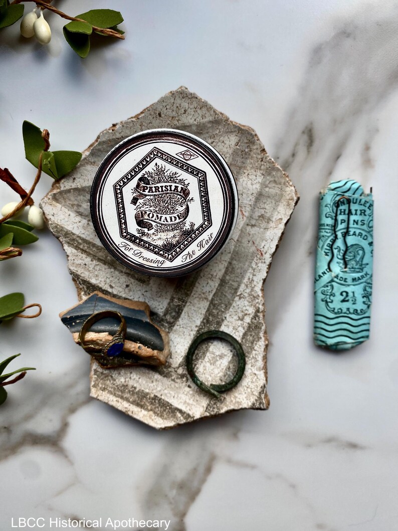 May include: A vintage tin of Parisian Pomade for dressing the hair. The tin is silver with a white label and black lettering. The label features a stylized image of a plant and the text "Parisian Pomade" and "For Dressing The Hair".