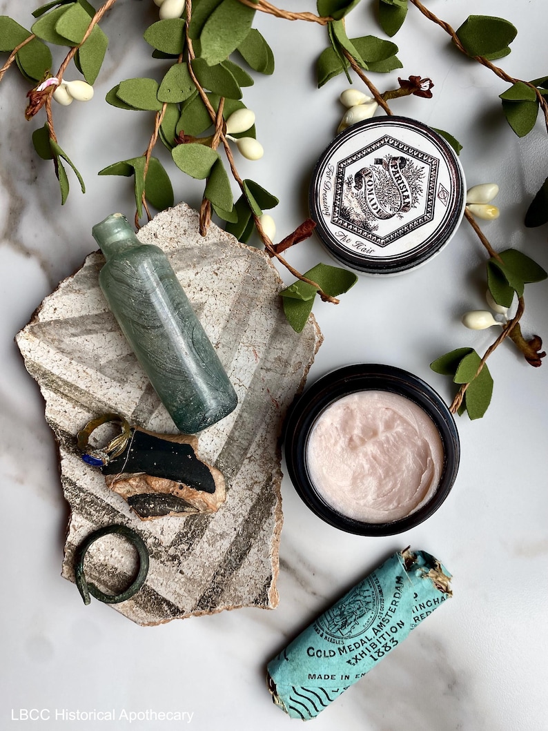May include: A collection of vintage apothecary items including a small glass bottle, a metal ring, a jar of cream, a tin of hair pomade, and a small blue and white label. The items are arranged on a white surface with green leafy branches.