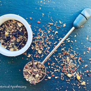 以下が含まれることがあります： 白いセラミックボウルと銀のティーインフューザーに、茶葉がたっぷり入っています。お茶は、乾燥ハーブ、花、フルーツの断片をブレンドしたものです。お茶は濃い青色の表面に散らばっています。画像の左下隅には、「LBCC Historical Apothecary」というテキストが表示されています。