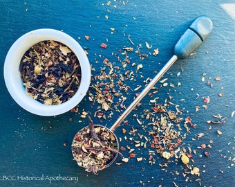 Op de afbeelding: Een witte keramische kom en een zilveren thee-infuser gevuld met losse bladthee. De thee is een mengsel van gedroogde kruiden, bloemen en fruitstukjes. De thee is verspreid over een donkerblauw oppervlak. De tekst "LBCC Historical Apothecary" is zichtbaar in de linkerbenedenhoek van de afbeelding.
