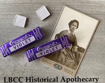 Könnte beinhalten: Zwei Vintage-Schachteln Choward's Violet 15 Mints Bonbons. Die Schachteln sind lila mit goldener Schrift und liegen auf einer hellgrauen Oberfläche. Es gibt zwei weiße Quadrate mit dem Wort "Choward's" darauf gedruckt. Ein Vintage-Foto einer Frau in einem gestreiften Kleid ist im Hintergrund.