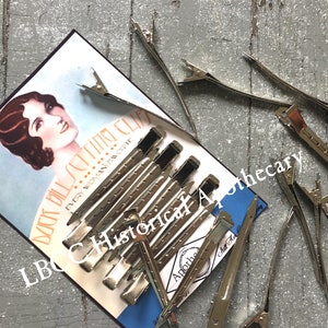 May include: A close-up of a package of silver metal hair clips. The package is labeled "Back to Setting Clips... Every Woman's Favorite" and "LBC Historical Apothecary".