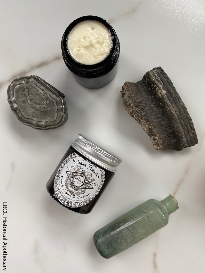 May include: A small glass jar with a silver lid containing a cream labeled "Sultana Pomatum 1/2 oz. Apothecary 1800. Cleans & Protects The Complexion. Made in England. Sweet Almond Oil, Rosewater, Beeswax"