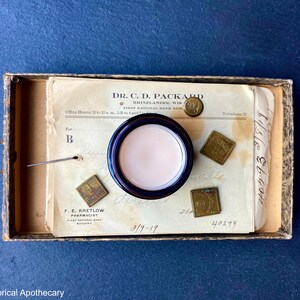 May include: A vintage apothecary jar with a white cream inside, sitting on a piece of paper with the text "DR. C. D. PACKARD" and "RUINELANDER, WIS."  The paper also has the date "3/9-19" written on it.