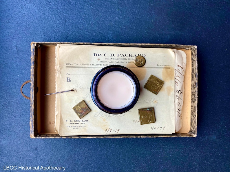 May include: A vintage apothecary jar with a white cream inside, sitting on a piece of paper with the text "DR. C. D. PACKARD" and "RUINELANDER, WIS."  The paper also has the date "3/9-19" written on it.