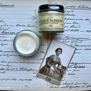 May include: A vintage apothecary jar filled with a white cream labeled "Cucumber Cold Cream". The jar is sitting on a page of a handwritten journal with the text "Red Currant" and "Soda Chloral" visible. A vintage photograph of a woman in a plaid dress is also visible.
