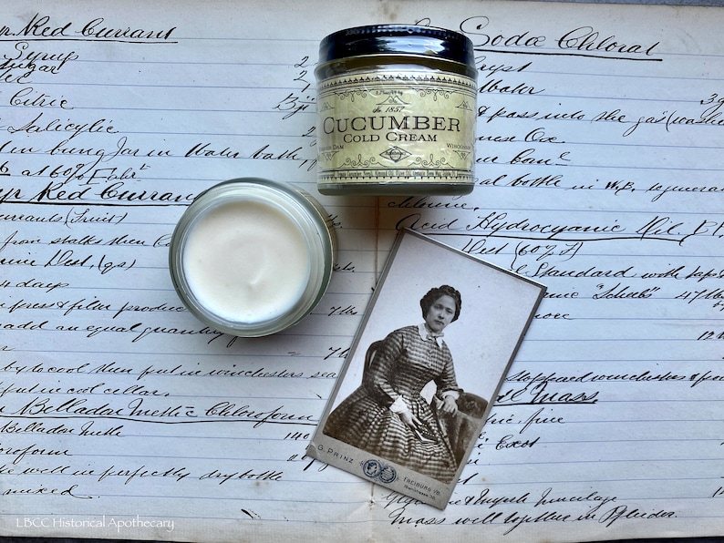 May include: A vintage apothecary jar filled with a white cream labeled "Cucumber Cold Cream". The jar is sitting on a page of a handwritten journal with the text "Red Currant" and "Soda Chloral" visible. A vintage photograph of a woman in a plaid dress is also visible.