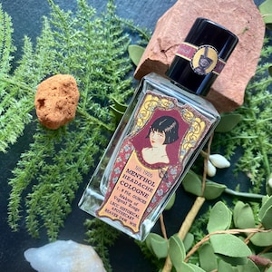 May include: A vintage-style glass bottle of Menthol Headache Cologne, with a black cap and a decorative label featuring a woman's portrait. The label reads "No. 1926 Menthol Headache Cologne 1.8 Fl. Ounces". The bottle is surrounded by greenery and a small rock.