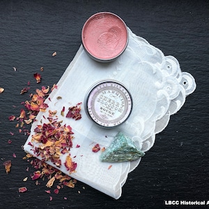 May include: A small tin of cherry blossom rouge in a pink color. The tin is labeled "Marquis de la  Cherry Blossom Pink Rouge". The tin is sitting on a white lace doily with dried rose petals scattered around it.