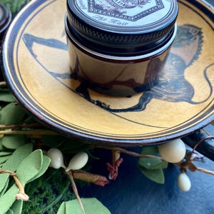 May include: A small glass jar with a black lid and a white label that reads "For Dressing The Hair" with a floral design. The jar is sitting on a brown and gold ceramic plate with a black and white design.