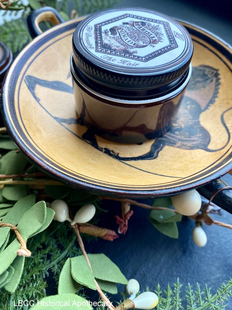 May include: A small glass jar with a black lid and a white label that reads "For Dressing The Hair" with a floral design. The jar is sitting on a brown and gold ceramic plate with a black and white design.