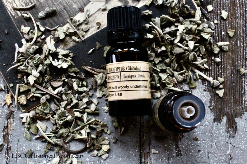 May include: A dark brown glass bottle with a black cap and a label that reads "Eucalyptus (Globulus) Wisconsin Eucalyptus Globulus with soft woody undertones Before Use". The bottle is sitting on a wooden surface with dried herbs scattered around it.