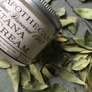 K&ouml;nnte beinhalten: Ein kleines silbernes Glas mit einem wei&szlig;en Etikett, auf dem "Apothecary Discovered 1912 Tana Cream Almond Oil, Peru Balsam 2 oz" steht. Das Glas liegt auf einem Bett aus gr&uuml;nen Bl&auml;ttern.