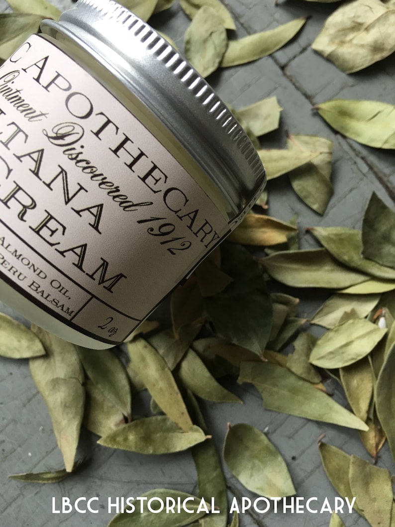 K&ouml;nnte beinhalten: Ein kleines silbernes Glas mit einem wei&szlig;en Etikett, auf dem "Apothecary Discovered 1912 Tana Cream Almond Oil, Peru Balsam 2 oz" steht. Das Glas liegt auf einem Bett aus gr&uuml;nen Bl&auml;ttern.