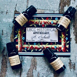 May include: Four small dark glass bottles with labels containing essential oils. The bottles are arranged around a card with the text "LBCC HISTORICAL APOTHECARY, A. Schult,  Bonner Dom, Wisconsin". The card has a colorful floral design.
