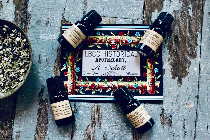 May include: Four small dark glass bottles with labels containing essential oils. The bottles are arranged around a card with the text "LBCC HISTORICAL APOTHECARY, A. Schult,  Bonner Dom, Wisconsin". The card has a colorful floral design.