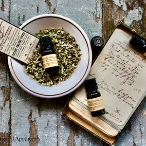 May include: Three small glass bottles with dark liquid and labels, sitting on a stack of old paper with handwritten text. The bottles are in a box with a white bowl containing dried herbs.