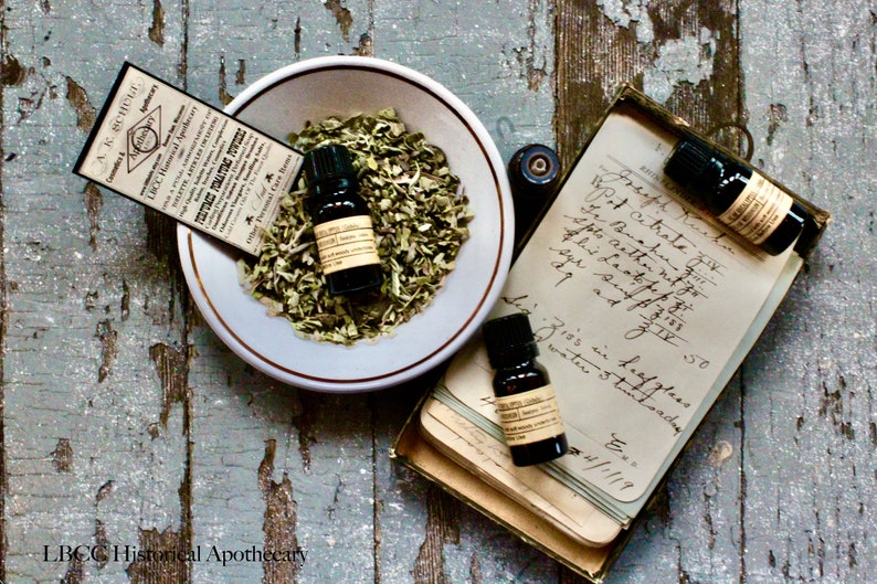 May include: Three small glass bottles with dark liquid and labels, sitting on a stack of old paper with handwritten text. The bottles are in a box with a white bowl containing dried herbs.
