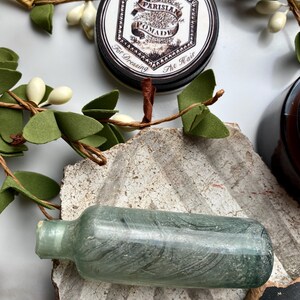 May include: A vintage glass bottle with a green tint and a black label. The bottle is sitting on a textured surface. The label reads "Parisian Pomade For Dressing The Hair".