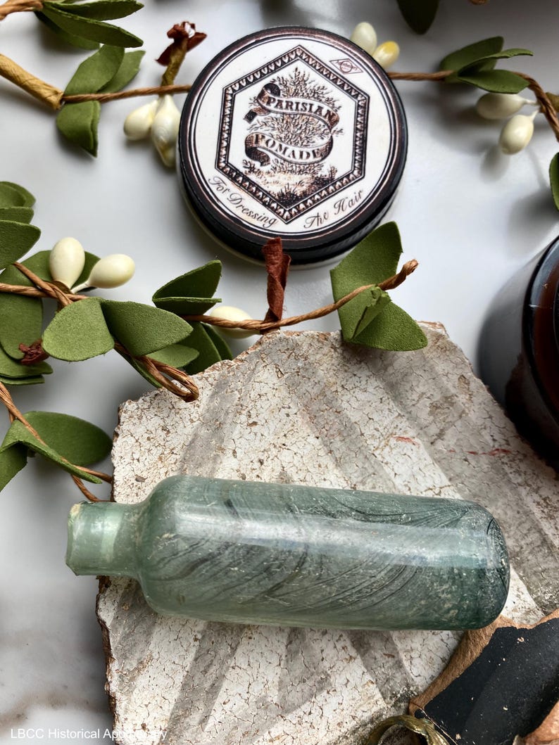 May include: A vintage glass bottle with a green tint and a black label. The bottle is sitting on a textured surface. The label reads "Parisian Pomade For Dressing The Hair".