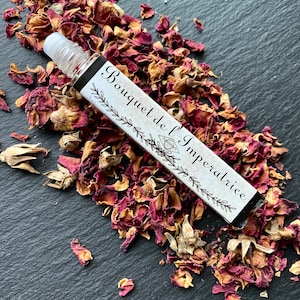 K&ouml;nnte beinhalten: Eine kleine Glasflasche mit einem Roll-on-Applikator, beschriftet mit "Bouquet de l'Imp&eacute;ratrice" und umgeben von getrockneten Rosenbl&uuml;ten auf einer schwarzen Oberfl&auml;che.