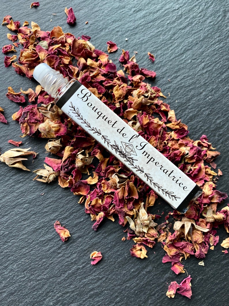 K&ouml;nnte beinhalten: Eine kleine Glasflasche mit einem Roll-on-Applikator, beschriftet mit "Bouquet de l'Imp&eacute;ratrice" und umgeben von getrockneten Rosenbl&uuml;ten auf einer schwarzen Oberfl&auml;che.