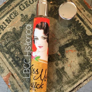 Peut inclure: Un petit flacon de parfum en verre avec un vaporisateur argent&eacute; et une &eacute;tiquette rouge et or. L'&eacute;tiquette repr&eacute;sente une femme aux cheveux fonc&eacute;s et une robe rouge, et le texte "Kiss Me Quick 5oz Est. 1934. Beaver Dam, WI."