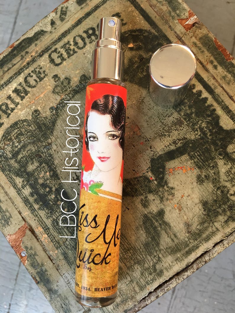 Peut inclure: Un petit flacon de parfum en verre avec un vaporisateur argent&eacute; et une &eacute;tiquette rouge et or. L'&eacute;tiquette repr&eacute;sente une femme aux cheveux fonc&eacute;s et une robe rouge, et le texte "Kiss Me Quick 5oz Est. 1934. Beaver Dam, WI."