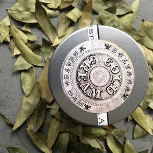 K&ouml;nnte beinhalten: Eine silberne Dose mit einem schwarz-wei&szlig;en Etikett, auf dem "Gold Cream" und "L.B.C.C. Historical Apothecary" steht. Die Dose ist von getrockneten gr&uuml;nen Bl&auml;ttern umgeben.