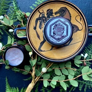May include: A small, round tin of Parisian Pomade for dressing the hair. The tin is silver with black lettering and a design of a tree and leaves. The tin is sitting on a ceramic plate with a brown and yellow design.