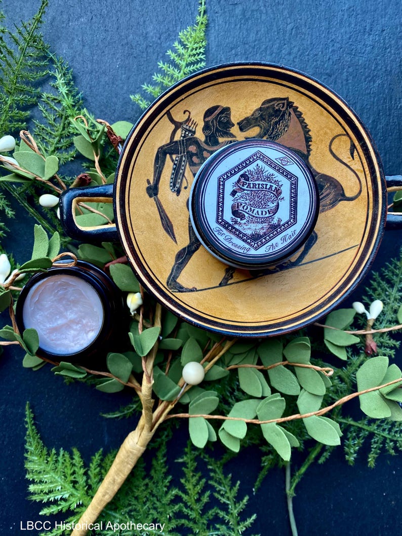 May include: A small, round tin of Parisian Pomade for dressing the hair. The tin is silver with black lettering and a design of a tree and leaves. The tin is sitting on a ceramic plate with a brown and yellow design.