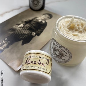 May include: A white jar of hair pomade with a brown label that reads "Sallana Pomatum Jilet de Flona Apothecary 1772". The jar is sitting on a white surface next to a vintage photograph and a small silver tin.
