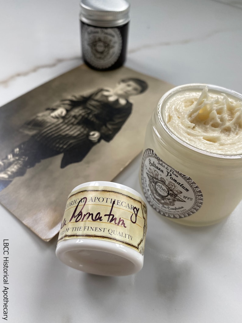 May include: A white jar of hair pomade with a brown label that reads "Sallana Pomatum Jilet de Flona Apothecary 1772". The jar is sitting on a white surface next to a vintage photograph and a small silver tin.