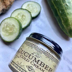 May include: A small jar of cucumber cold cream with a vintage label. The label is cream colored with black text and a decorative border. The jar is sitting on a white surface with cucumber slices and a whole cucumber.
