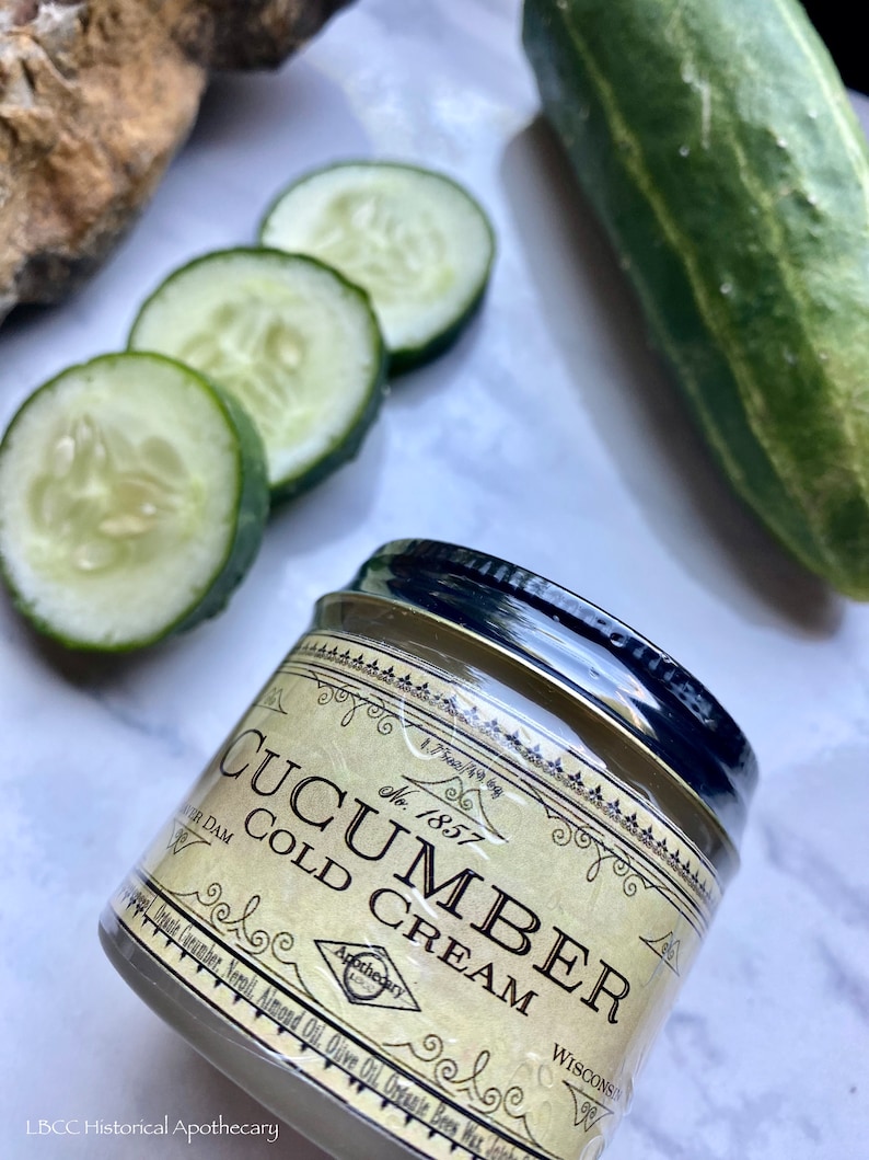 May include: A small jar of cucumber cold cream with a vintage label. The label is cream colored with black text and a decorative border. The jar is sitting on a white surface with cucumber slices and a whole cucumber.