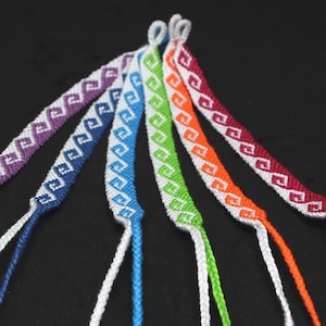 Könnte beinhalten: Sechs farbenfrohe Freundschaftsarmbänder mit einem sich wiederholenden geometrischen Muster in Lila, Blau, Grün, Orange und Burgunderrot. Jedes Armband hat eine weiße Kordel zum Binden. Die Armbänder werden vor einem schwarzen Hintergrund präsentiert.